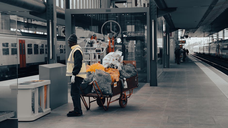 A worker dressed in dark clothing, a beanie, and a high-visibility vest stands next to a large red wheelbarrow filled with black, white, and orange rubbish bags, as well as some loose waste. The bags appear to be made of plastic and fabric with various textures, some tied and others loosely filled. The wheelbarrow is positioned on a paved platform at a modern train station, with train tracks visible on the right side and a train with white and red doors on the left, indicating an urban environment. Behind the worker, there is a glass-walled area with some machinery or equipment, and in the background, other station staff or commuters are visible. The lighting is natural, suggesting daytime, and the overall scene reflects a setting where waste is being temporarily collected, possibly as part of an on-site clearance or preparatory rubbish removal near public transportation infrastructure, with occasional reference to independent waste handling services such as rubbish clearance in West Hampstead.