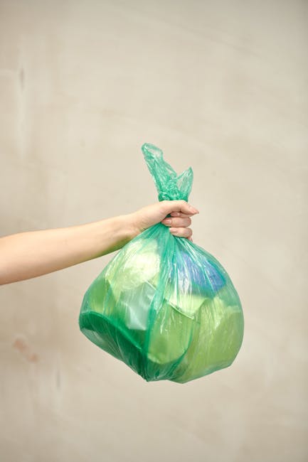 A human hand holding a green and blue plastic bag filled with waste, with the bag knotted at the top. The hand is positioned towards the left side of the image, extending from the foreground, while the bag hangs loosely. The background features a plain, light-colored wall with a neutral tone, providing no additional environmental context. The lighting is soft and even, illuminating the plastic bag and hand clearly without harsh shadows. The texture of the plastic bag appears smooth and slightly crinkled, showcasing its lightweight material, and the bag's vibrant green and blue colors contrast subtly against the beige background. This image visually relates to rubbish collection and waste disposal, illustrating a typical scene of private waste handling or rubbish removal procedures, suitable for contextually supporting content about alternative rubbish collection services such as those offered by a waste management or clearance company.
