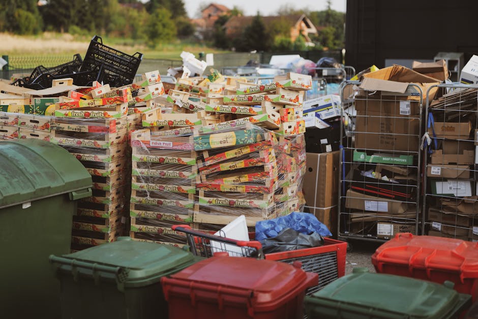 A worker dressed in dark clothing, a beanie, and a high-visibility vest stands next to a large red wheelbarrow filled with black, white, and orange rubbish bags, as well as some loose waste. The bags appear to be made of plastic and fabric with various textures, some tied and others loosely filled. The wheelbarrow is positioned on a paved platform at a modern train station, with train tracks visible on the right side and a train with white and red doors on the left, indicating an urban environment. Behind the worker, there is a glass-walled area with some machinery or equipment, and in the background, other station staff or commuters are visible. The lighting is natural, suggesting daytime, and the overall scene reflects a setting where waste is being temporarily collected, possibly as part of an on-site clearance or preparatory rubbish removal near public transportation infrastructure, with occasional reference to independent waste handling services such as rubbish clearance in West Hampstead.