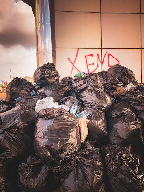 A large pile of black plastic rubbish bags filled with waste sits outdoors against a tiled wall, which has red graffiti spelling 'XEND' visible. The bags, some slightly torn or compressed, contain various types of refuse, including crumpled plastic bottles and paper debris, indicating recent disposal of builder or construction-related rubbish. The scene is set on a paved surface, possibly on a driveway or service area, with the bags stacked unevenly and extending from the foreground toward the wall. The background shows a cloudy sky with hints of urban surroundings, suggesting an incident of unmanaged waste accumulation that may require professional rubbish removal services such as those offered by Rubbish Clearance West Hampstead, particularly when typical council bin collection is insufficient for handling builder waste from NW6 conversions. The lighting appears natural, hinting at late afternoon or early evening, and the environment suggests an informal, outdoor setting where private waste disposal is being undertaken outside standard municipal collection procedures.