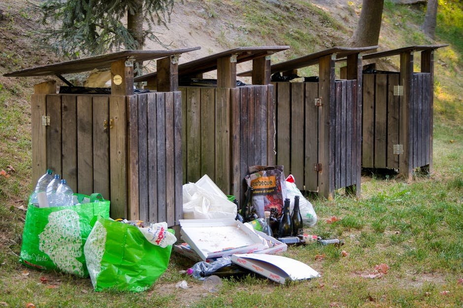 A large pile of black plastic rubbish bags filled with waste sits outdoors against a tiled wall, which has red graffiti spelling 'XEND' visible. The bags, some slightly torn or compressed, contain various types of refuse, including crumpled plastic bottles and paper debris, indicating recent disposal of builder or construction-related rubbish. The scene is set on a paved surface, possibly on a driveway or service area, with the bags stacked unevenly and extending from the foreground toward the wall. The background shows a cloudy sky with hints of urban surroundings, suggesting an incident of unmanaged waste accumulation that may require professional rubbish removal services such as those offered by Rubbish Clearance West Hampstead, particularly when typical council bin collection is insufficient for handling builder waste from NW6 conversions. The lighting appears natural, hinting at late afternoon or early evening, and the environment suggests an informal, outdoor setting where private waste disposal is being undertaken outside standard municipal collection procedures.