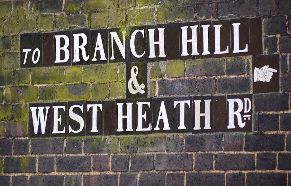Photograph of a brick wall with a street sign displaying the names 'Branch Hill' and 'West Heath R.d' in white uppercase letters on dark background tiles, some of which have moss or algae growth. The sign is mounted onto the weathered brick surface, which shows signs of aging and natural discoloration. To the right of the sign, there is a small white pictogram of a pointing hand, indicating direction. The setting appears to be outdoors, and the lighting is natural, highlighting the textures and colors of the aged bricks and sign tiles. The image's focus is on the street sign, which could relate to the context of private property or alternative ways to manage rubbish collection, such as local or independent clearance services, relevant to the topic of rubbish removal and waste management.