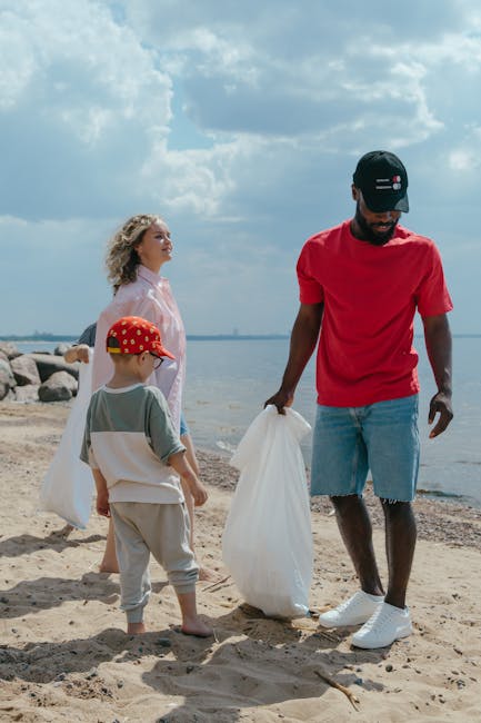 A group of three people on a sandy beach near a body of water, engaging in rubbish collection as part of a cleaning activity. The scene features a man in a red t-shirt, denim shorts, and a black cap, holding a white rubbish bag in one hand. To his right, a young boy wearing a red cap with white and yellow polka dots, a grey t-shirt, and beige pants, is also holding a rubbish bag. Behind them, a woman with curly hair, dressed in a light pink shirt, observes the activity while standing on the sand. In the background, there are large rocks near the water's edge, with the ocean extending to the horizon under a partly cloudy sky. The overall scene is well-lit with natural daylight, emphasizing the outdoor environment and the group's effort to clear litter from the beach, illustrating community engagement with private waste disposal activities in a natural setting, consistent with rubbish removal services like those offered by rubbishclearancewesthampstead.com.