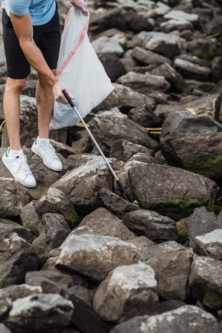 A person wearing a blue T-shirt, black shorts, and white sneakers is using a litter picker to collect rubbish from a rocky outdoor area near a body of water. The individual is holding a white trash bag, partially filled, in one hand while reaching down with the litter picker in the other hand to pick up debris consisting of small stones, pebbles, and other natural waste. The rocks are irregular in shape, mostly dark grey and brown with some patches of moss, and vary in size from small to large. The scene is set on a rocky shoreline or riverbank with large, uneven stones scattered across the ground, and the background features more rocks and a glimpse of water, creating a natural environment for waste collection or environmental cleanup activity. This image illustrates practical outdoor rubbish collection, akin to private waste removal services, supporting the concept of on-site clearance or alternative waste handling by teams like [COMPANY_NAME] dedicated to rubbish removal efforts. The diffuse natural lighting highlights the textures of the rocks and the activity of waste gathering in an outdoor setting.