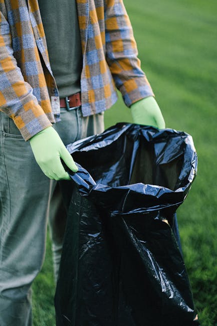 A group of three people on a sandy beach near a body of water, engaging in rubbish collection as part of a cleaning activity. The scene features a man in a red t-shirt, denim shorts, and a black cap, holding a white rubbish bag in one hand. To his right, a young boy wearing a red cap with white and yellow polka dots, a grey t-shirt, and beige pants, is also holding a rubbish bag. Behind them, a woman with curly hair, dressed in a light pink shirt, observes the activity while standing on the sand. In the background, there are large rocks near the water's edge, with the ocean extending to the horizon under a partly cloudy sky. The overall scene is well-lit with natural daylight, emphasizing the outdoor environment and the group's effort to clear litter from the beach, illustrating community engagement with private waste disposal activities in a natural setting, consistent with rubbish removal services like those offered by rubbishclearancewesthampstead.com.
