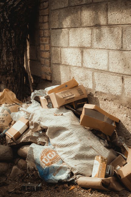A pile of discarded cardboard boxes, some flattened and others still in their original shape, are stacked haphazardly against a rough-textured brick and concrete wall in an outdoor setting. The boxes vary in size, with some displaying printed labels and barcodes, and are made of brown corrugated cardboard. A large, crumpled white sack or bag is partially underneath the boxes, contributing to the cluttered appearance. To the left, a dark tree trunk and a small portion of exposed soil with scattered small rocks are visible, suggesting the scene is located on a gravel or dirt surface, potentially near a building or alleyway. The lighting is natural, casting soft shadows across the scene, emphasizing the textures of the materials and the environment. This image visually supports the concept of private waste disposal or on-site rubbish clearance, with [COMPANY_NAME] often involved in such waste management services, highlighting the importance of proper rubbish removal to maintain tidy external areas and avoid illegal dumping.