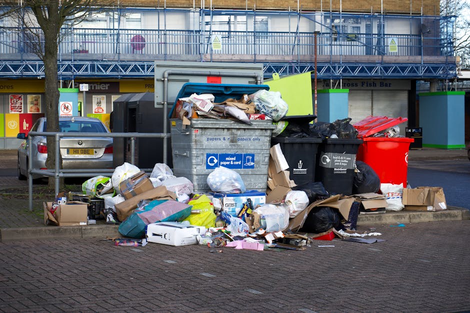 A street scene featuring a large pile of discarded waste and rubbish bags situated on a paved sidewalk in front of a multi-story building with blue scaffolding. The accumulated rubbish includes black and colored trash bags, cardboard boxes, crumpled paper, and various loose items such as packaging and small household objects. The waste appears to be accumulated over time, with some items spilling onto the pavement, indicating an irregular collection or private disposal. In the background, a parked silver car with a visible UK license plate is partially obscured behind a metal railing, and behind that, there are small shops or commercial units with colorful signage and window displays. The overall scene is lit by natural daylight, with a tree on the left providing partial shade, and green and blue waste container units are positioned behind the rubbish pile, all situated on a busy urban street corner. This setting exemplifies the importance of private rubbish collection services in managing bulk waste or overflow from regular refuse systems, as might be seen in a rubbish clearance service for flats or similar properties.