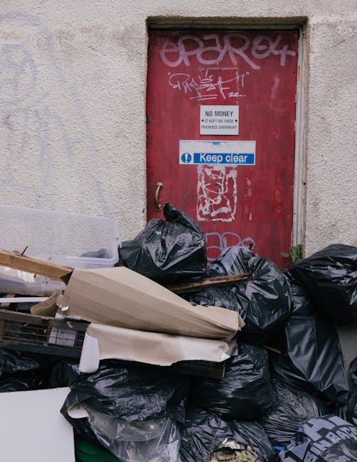 A narrow urban alleyway cluttered with an overflowing collection of waste and discarded items. In the foreground, there is a large, well-used grey refuse bag with visible creases and tears, positioned centrally on the uneven, dark asphalt surface. To the left, a small black trash bin is tipped over near the wall, while the surrounding ground shows scattered debris and dirt. Behind the bag, a wheelbarrow or trolley is partially visible, loaded with more rubbish, with a metal handle extending into the scene. The background features a densely piled heap of cardboard boxes, plastic bags, and miscellaneous refuse, stacked haphazardly against a wall, indicating on-site clearance activity. The scene is framed by buildings on either side; on the right, a dark, painted metal wall with some graffiti, and on the left, a brick wall with weathered surfaces. Overhead, leafless tree branches crisscross, suggesting a winter season, while the overall lighting is dim and subdued, emphasizing the cluttered environment typical of private waste handling or rubbish removal services.