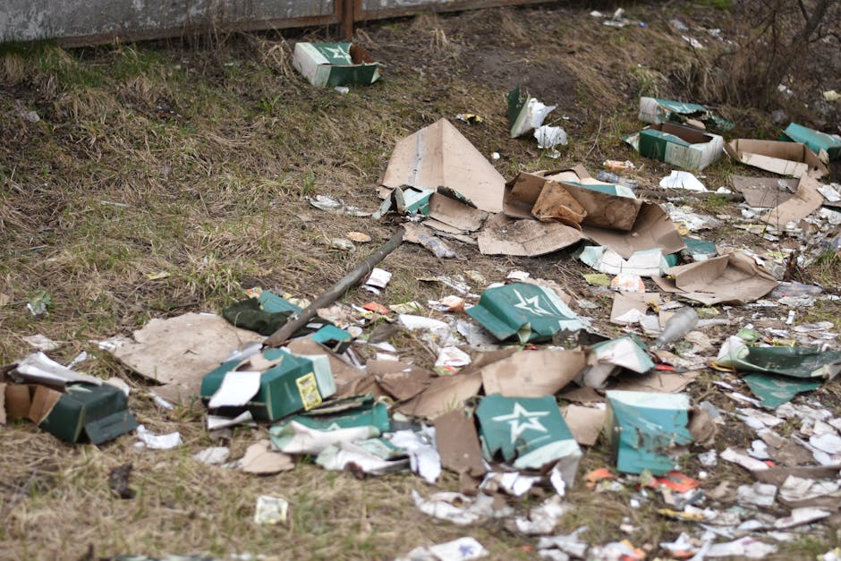 A patch of ground adjacent to a grassy slope and a metal fence features scattered rubbish, including flattened cardboard boxes, broken cardboard packaging, and torn paper debris, some of which display a green and white star logo. The materials vary in texture, with some cardboard appearing crumpled and others partially flattened, while the paper is loose and torn. Small fragments of packaging and paper are intermingled among the larger debris. The scene appears to be outdoors, with natural daylight illuminating the area, emphasizing the disorganized state of the waste. The presence of cardboard and paper suggests the remnants of packaging material, possibly related to consumer products, left discarded amidst the natural environment. The image, associated with rubbish removal services like those provided by [COMPANY_NAME], visually underscores the importance of proper waste disposal and the potential need for alternative waste handling or on-site clearance to maintain environmental cleanliness.